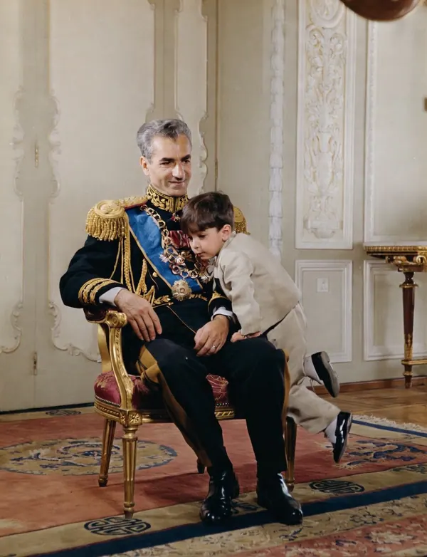 His Majesty, the Shah of Iran in a formal military uniform sits in a chair while a young boy leans against him — a public domain photograph from The Shah of Iran archive documenting the Pahlavi era.