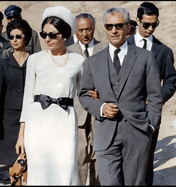 His Majesty, the Shah of Iran in a grey suit and Farah Pahlavi, Empress of Iran, in a white dress walk arm in arm outdoors — a public domain photograph from The Shah of Iran archive documenting the Pahlavi era.
