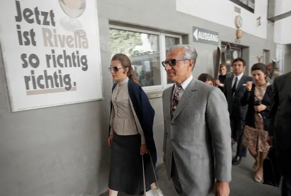 His Majesty, the Shah of Iran in a grey suit and Farah Pahlavi, Empress of Iran, in a dark cardigan walk past a sign — a public domain photograph from The Shah of Iran archive documenting the Pahlavi era.