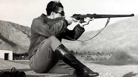 His Majesty, the Shah of Iran in a jacket and boots sits outdoors while aiming a rifle toward the distance — a public domain photograph from The Shah of Iran archive documenting the Pahlavi era.