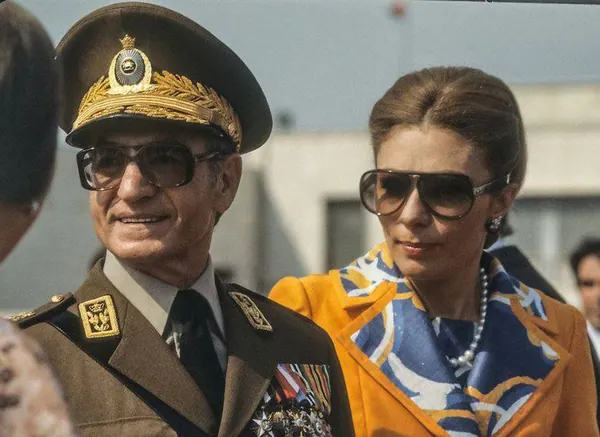 His Majesty, the Shah of Iran in a military uniform and Farah Pahlavi, Empress of Iran, in a yellow jacket wear sunglasses and smile — a public domain photograph from The Shah of Iran archive documenting the Pahlavi era.