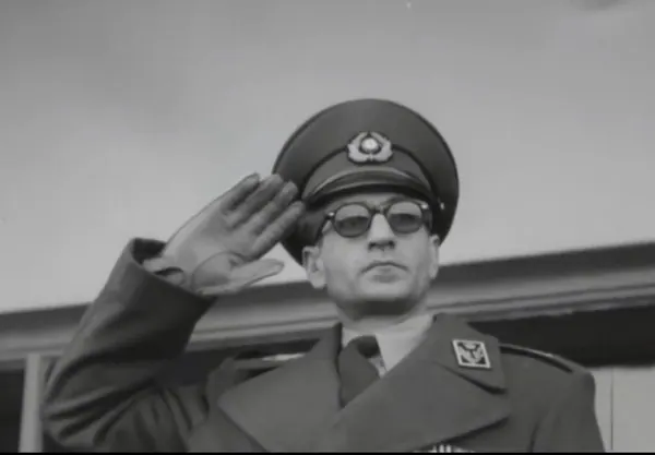His Majesty, the Shah of Iran in a military uniform and dark glasses salutes while wearing a peaked cap — a public domain photograph from The Shah of Iran archive documenting the Pahlavi era.