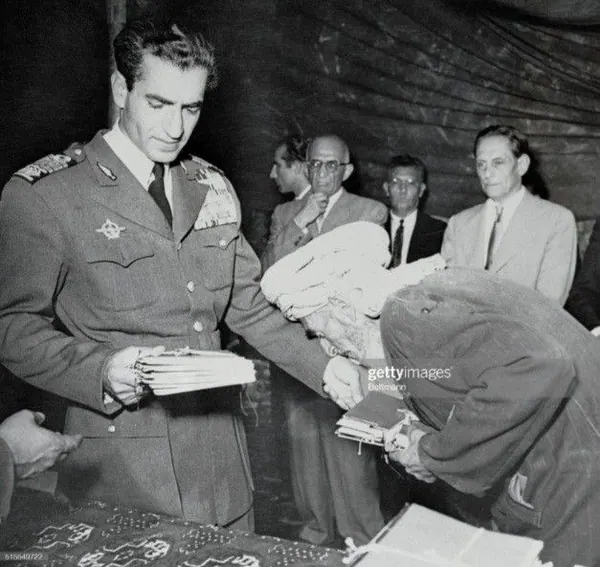 His Majesty, the Shah of Iran in a military uniform hands a stack of papers to a person bowing low — a public domain photograph from The Shah of Iran archive documenting the Pahlavi era.
