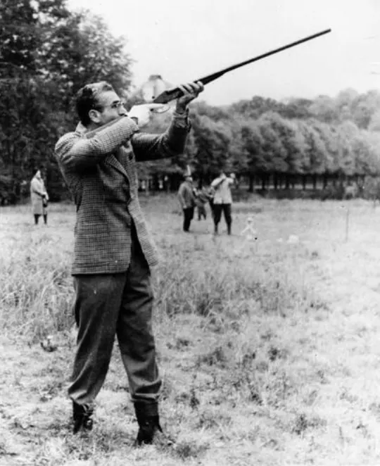 His Majesty, the Shah of Iran in a patterned jacket and trousers aims a long gun in an open grassy field — a public domain photograph from The Shah of Iran archive documenting the Pahlavi era.
