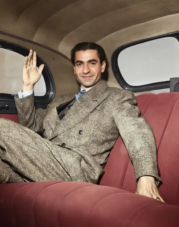 His Majesty, the Shah of Iran in a patterned suit sits in a red car seat and waves his hand — a public domain photograph from The Shah of Iran archive documenting the Pahlavi era.