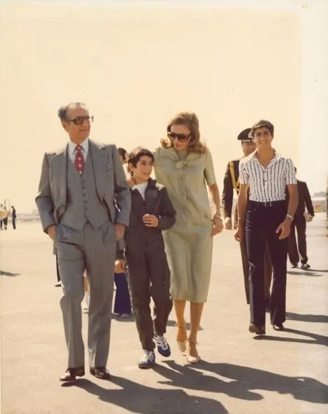 His Majesty, the Shah of Iran in a suit, Farah Pahlavi, Empress of Iran, in a dress, and two boys walk across a paved airfield — a public domain photograph from The Shah of Iran archive documenting the Pahlavi era.