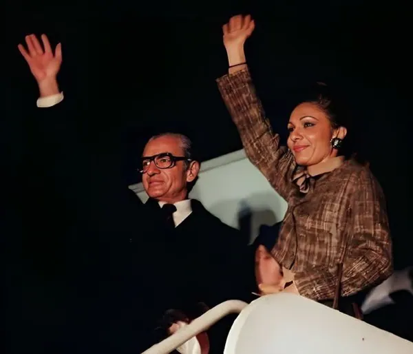 His Majesty, the Shah of Iran in a suit and Farah Pahlavi, Empress of Iran, in a patterned blouse wave their hands while standing together — a public domain photograph from The Shah of Iran archive documenting the Pahlavi era.