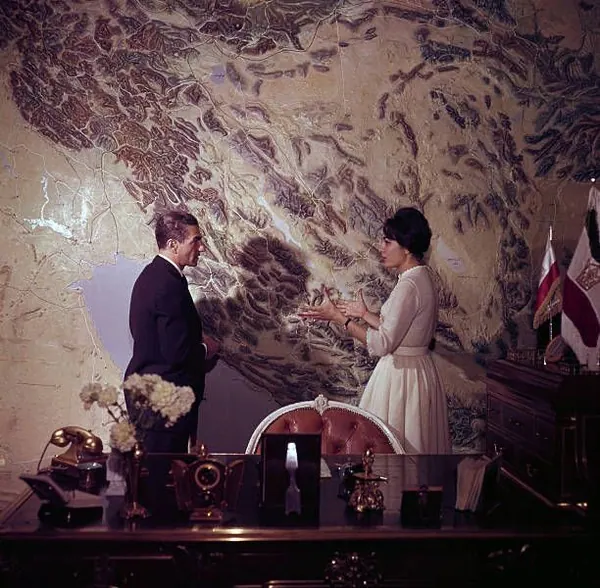 His Majesty, the Shah of Iran in a suit and Farah Pahlavi, Empress of Iran, in a white dress stand before a large wall map — a public domain photograph from The Shah of Iran archive documenting the Pahlavi era.