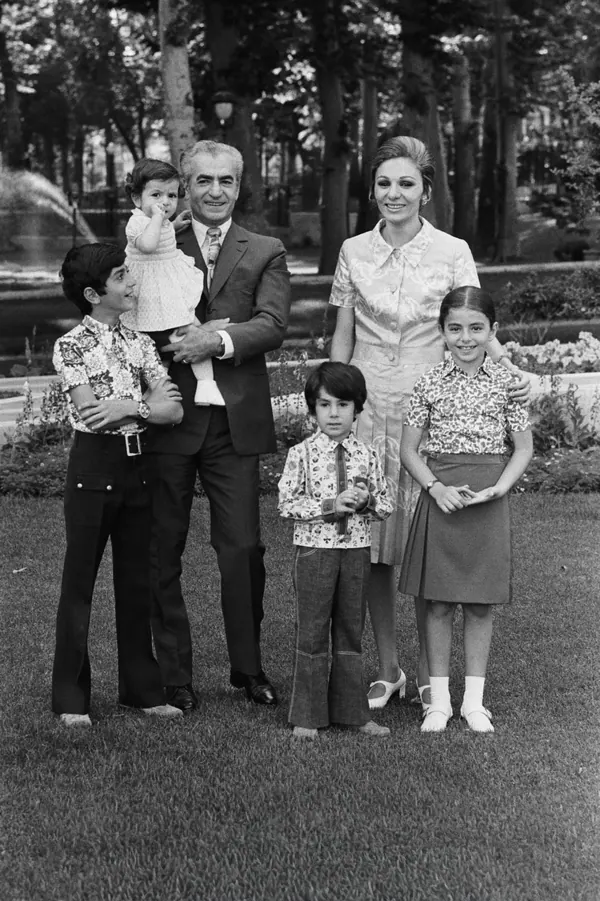 His Majesty, the Shah of Iran in a suit and Farah Pahlavi, Empress of Iran, in a dress stand with three children on a grassy lawn — a public domain photograph from The Shah of Iran archive documenting the Pahlavi era.