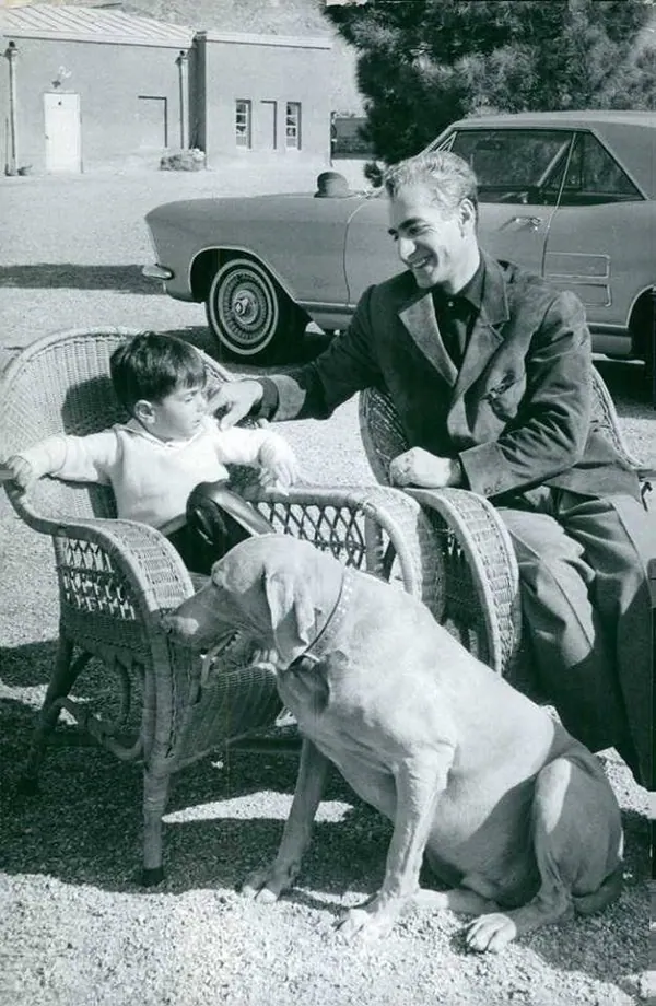 His Majesty, the Shah of Iran in a suit and a young child sit in wicker chairs beside a dog and car — a public domain photograph from The Shah of Iran archive documenting the Pahlavi era.