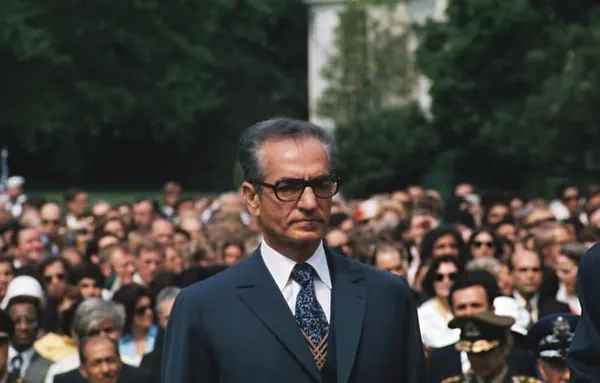 His Majesty, the Shah of Iran in a suit and glasses stands before a dense crowd of people outdoors — a public domain photograph from The Shah of Iran archive documenting the Pahlavi era.