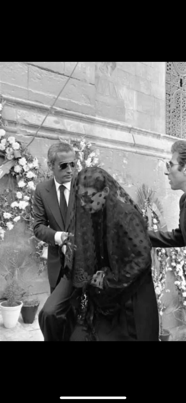 His Majesty, the Shah of Iran in a suit and sunglasses stands beside Farah Pahlavi, Empress of Iran, wearing a dark patterned veil and dress — a public domain photograph from The Shah of Iran archive documenting the Pahlavi era.