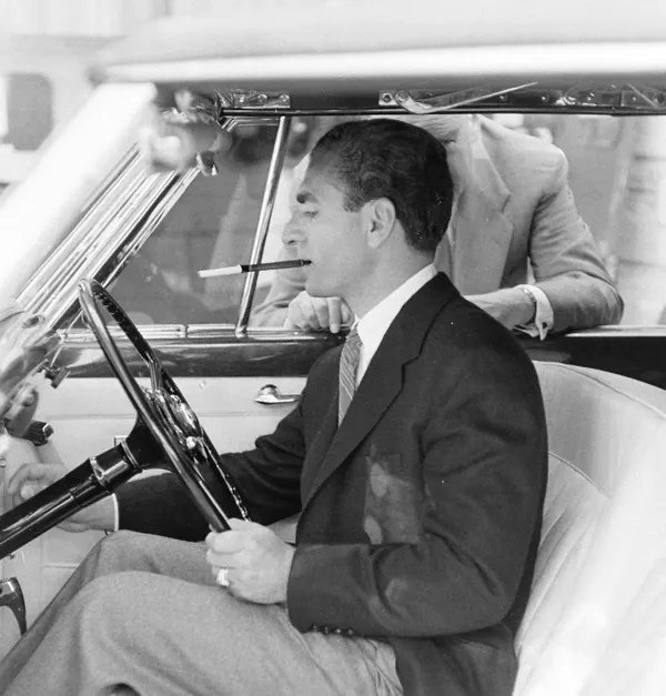 His Majesty, the Shah of Iran in a suit and tie smokes a cigarette while sitting in the driver's seat of a car — a public domain photograph from The Shah of Iran archive documenting the Pahlavi era.