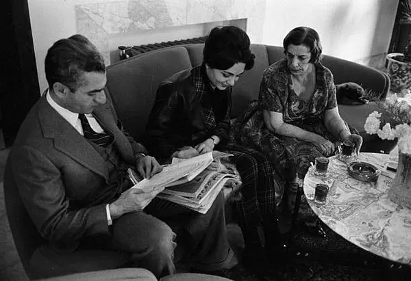 His Majesty, the Shah of Iran in a suit and two women sit on a sofa reading a newspaper together indoors — a public domain photograph from The Shah of Iran archive documenting the Pahlavi era.