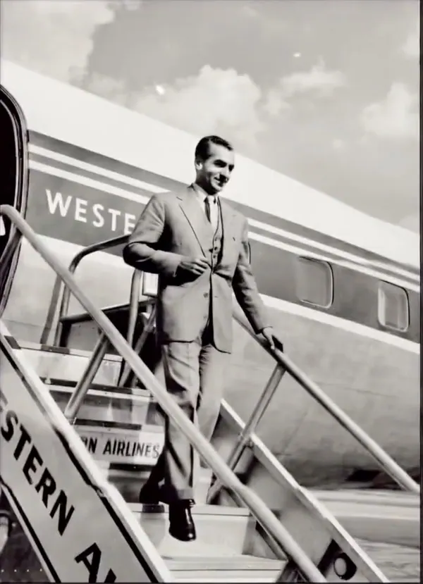 His Majesty, the Shah of Iran in a suit descends the stairs of a large airplane under a cloudy sky — a public domain photograph from The Shah of Iran archive documenting the Pahlavi era.