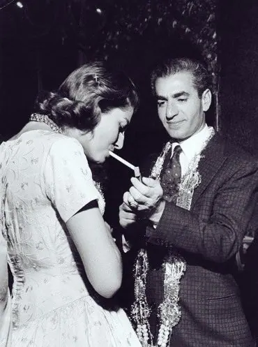 His Majesty, the Shah of Iran in a suit lights a cigarette for Farah Pahlavi, Empress of Iran, wearing a light-colored dress — a public domain photograph from The Shah of Iran archive documenting the Pahlavi era.