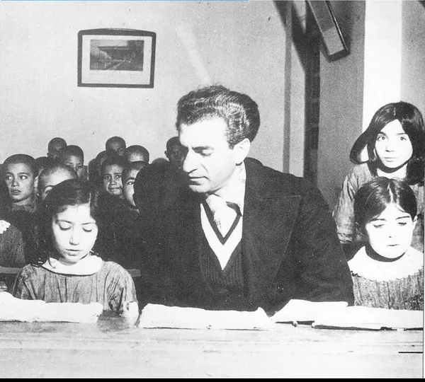 His Majesty, the Shah of Iran in a suit sits at a table with children in a room with a framed picture — a public domain photograph from The Shah of Iran archive documenting the Pahlavi era.
