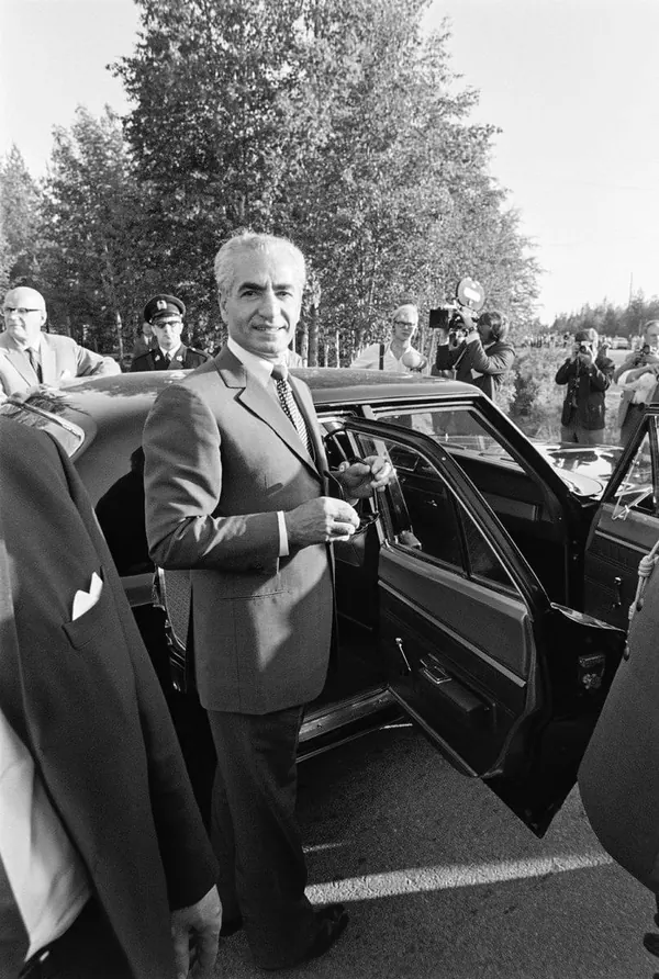 His Majesty, the Shah of Iran in a suit stands by an open car door while others watch in the background — a public domain photograph from The Shah of Iran archive documenting the Pahlavi era.