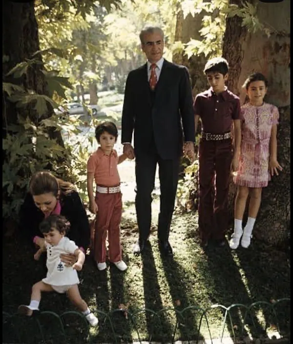 His Majesty, the Shah of Iran in a suit stands with four children in a wooded area near a low fence — a public domain photograph from The Shah of Iran archive documenting the Pahlavi era.