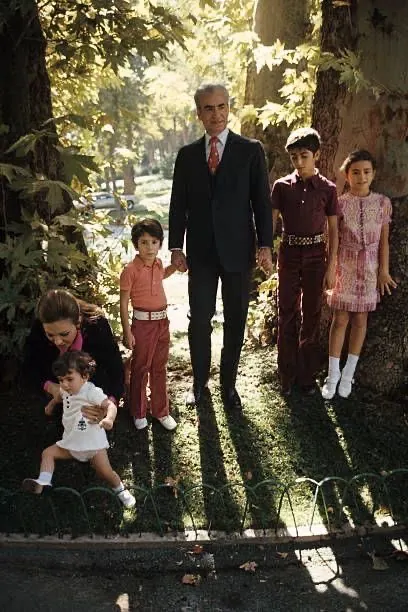 His Majesty, the Shah of Iran in a suit stands with three children in a wooded area while Farah Pahlavi, Empress of Iran, sits nearby — a public domain photograph from The Shah of Iran archive documenting the Pahlavi era.