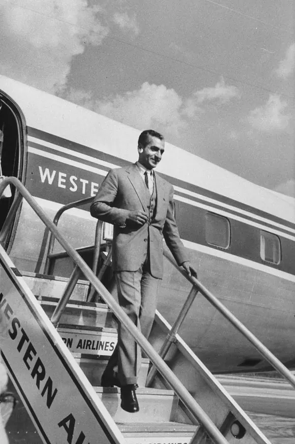 His Majesty, the Shah of Iran in a suit walks down the stairs of a large airplane on a tarmac — a public domain photograph from The Shah of Iran archive documenting the Pahlavi era.