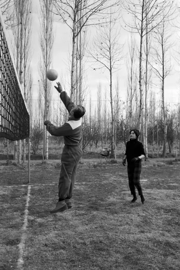 His Majesty, the Shah of Iran in a tracksuit jumps to hit a ball over a net while Farah Pahlavi, Empress of Iran, watches nearby — a public domain photograph from The Shah of Iran archive documenting the Pahlavi era.