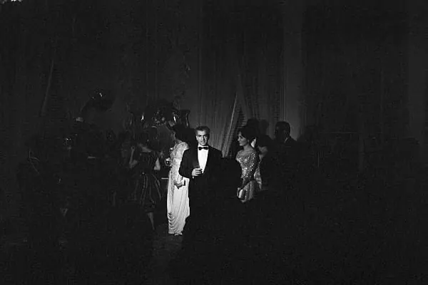 His Majesty, the Shah of Iran in a tuxedo and women in evening gowns stand in a dimly lit room with curtains — a public domain photograph from The Shah of Iran archive documenting the Pahlavi era.