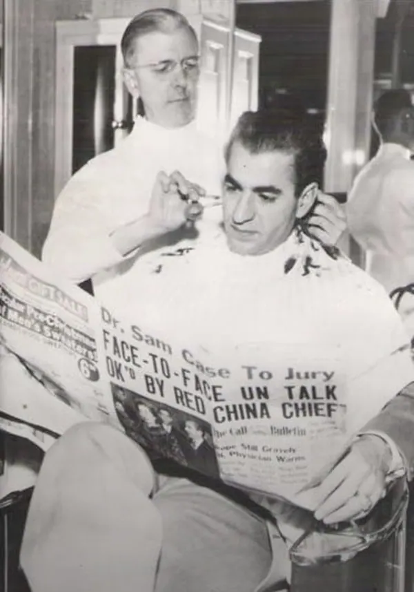 His Majesty, the Shah of Iran in a white cape reads a newspaper while a barber trims his sideburns with a razor — a public domain photograph from The Shah of Iran archive documenting the Pahlavi era.
