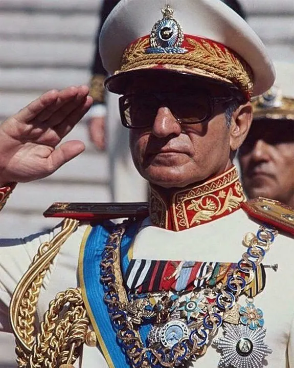 His Majesty, the Shah of Iran in a white military uniform with medals and sunglasses salutes while wearing a peaked cap — a public domain photograph from The Shah of Iran archive documenting the Pahlavi era.