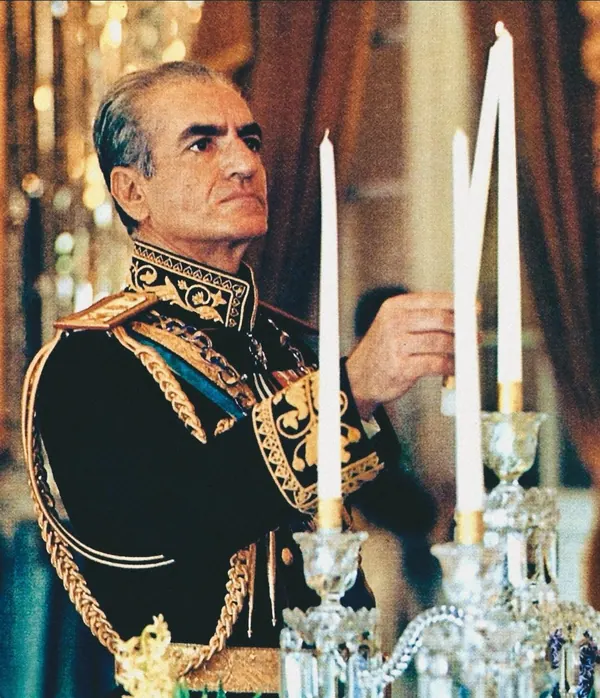 His Majesty, the Shah of Iran in an ornate dark military uniform with gold embroidery stands beside tall white candles — a public domain photograph from The Shah of Iran archive documenting the Pahlavi era.