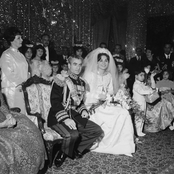 His Majesty, the Shah of Iran in military uniform and Farah Pahlavi, Empress of Iran, in a wedding dress sit together surrounded by guests — a public domain photograph from The Shah of Iran archive documenting the Pahlavi era.