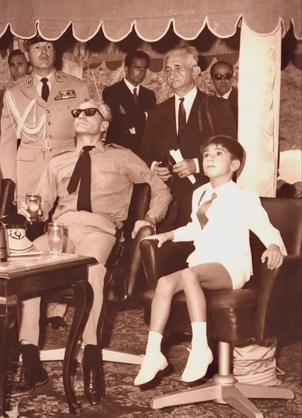 His Majesty, the Shah of Iran in sunglasses and a boy in white sit while men in suits and uniform stand behind — a public domain photograph from The Shah of Iran archive documenting the Pahlavi era.