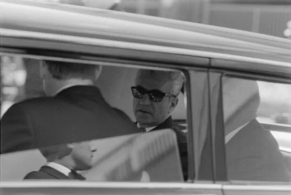 His Majesty, the Shah of Iran wearing dark sunglasses and a suit looks out from the back seat of a car — a public domain photograph from The Shah of Iran archive documenting the Pahlavi era.