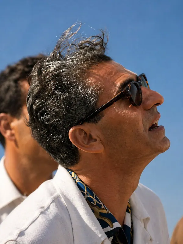 His Majesty, the Shah of Iran with curly hair and sunglasses looks upward while wearing a white jacket and patterned shirt — a public domain photograph from The Shah of Iran archive documenting the Pahlavi era.