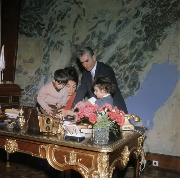 His Majesty, the Shah of Iran, woman, and two young children gather around a large ornate desk in front of a map — a public domain photograph from The Shah of Iran archive documenting the Pahlavi era.