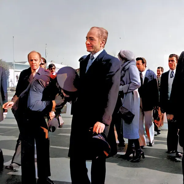 Men in dark suits and Farah Pahlavi, Empress of Iran, in a light coat stand outdoors on a paved surface — a public domain photograph from The Shah of Iran archive documenting the Pahlavi era.