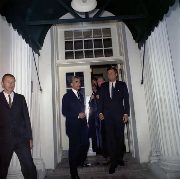 Men in dark suits walk through a white doorway under a dark canopy between two white columns — a public domain photograph from The Shah of Iran archive documenting the Pahlavi era.