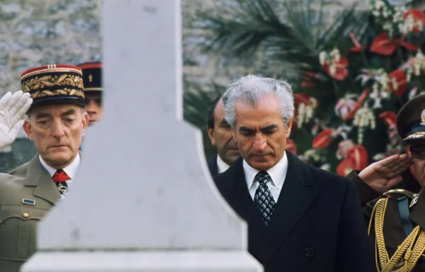 Men in military uniforms and formal suits stand solemnly beside a stone monument and floral arrangements — a public domain photograph from The Shah of Iran archive documenting the Pahlavi era.