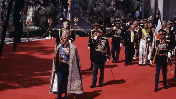 Men in ornate military uniforms and formal attire walk across a wide red carpet outdoors — a public domain photograph from The Shah of Iran archive documenting the Pahlavi era.