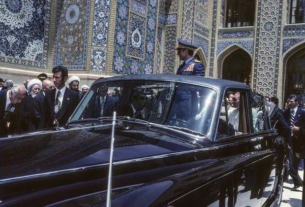Men in suits and uniforms stand beside a black luxury car in front of a tiled wall — a public domain photograph from The Shah of Iran archive documenting the Pahlavi era.