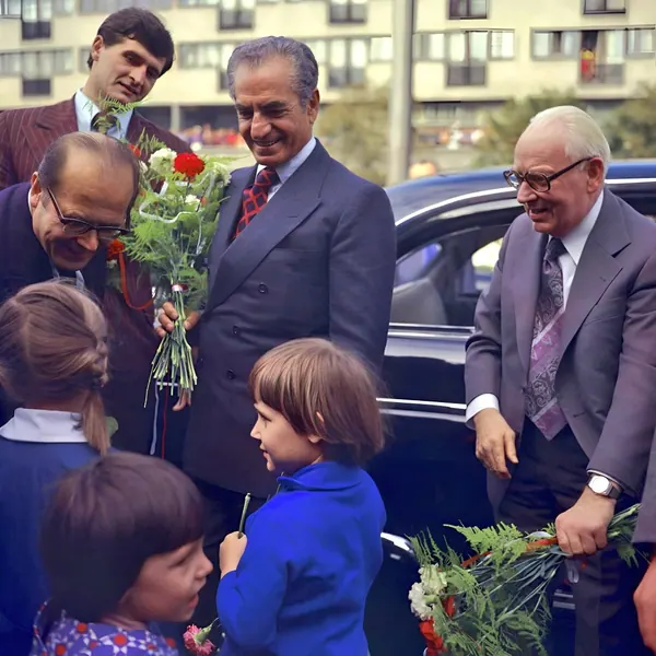 Men in suits stand by a black car while children hold flowers in an outdoor urban setting — a public domain photograph from The Shah of Iran archive documenting the Pahlavi era.