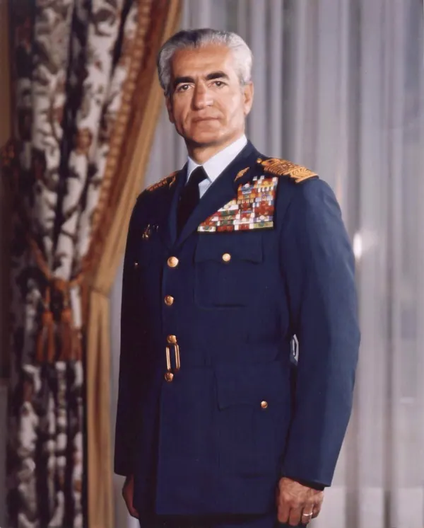 His Majesty, the Shah of Iran in a blue military uniform with medals stands before a curtained background — a public domain photograph from The Shah of Iran archive documenting the Pahlavi era.