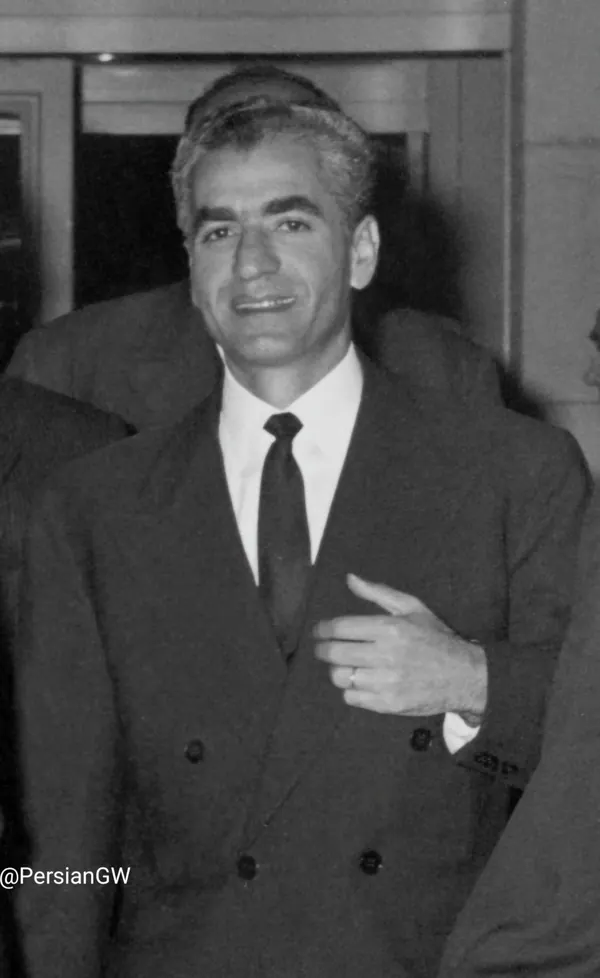 His Majesty, the Shah of Iran in a dark double-breasted suit and tie smiles while gesturing with his hand — a public domain photograph from The Shah of Iran archive documenting the Pahlavi era.