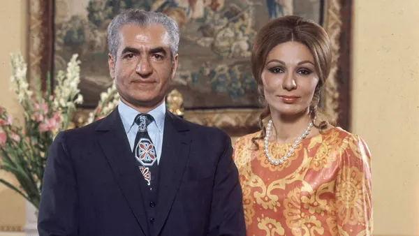 His Majesty, the Shah of Iran in a dark suit and Farah Pahlavi, Empress of Iran, in a patterned orange dress pose together — a public domain photograph from The Shah of Iran archive documenting the Pahlavi era.