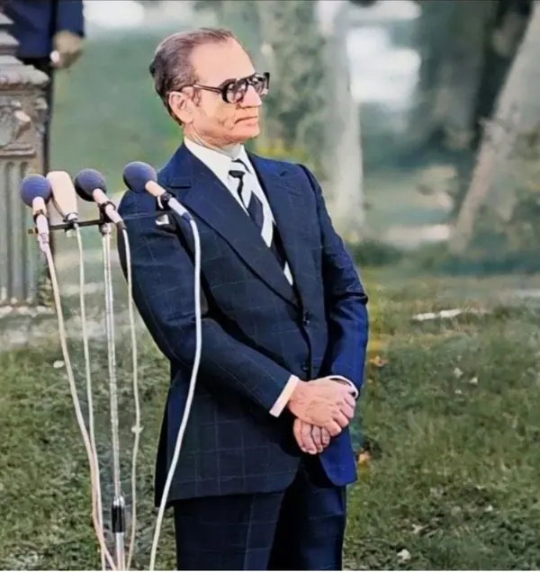 His Majesty, the Shah of Iran in a dark suit and glasses stands behind three microphones in an outdoor setting — a public domain photograph from The Shah of Iran archive documenting the Pahlavi era.