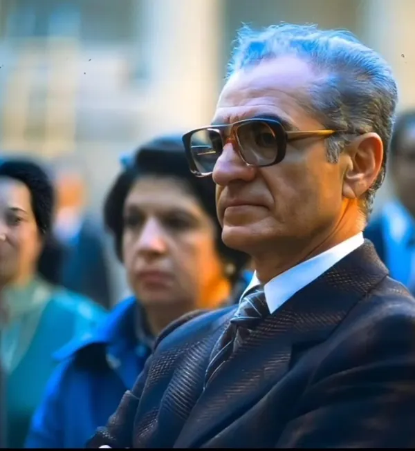 His Majesty, the Shah of Iran in a dark suit and glasses looks off-camera with two people blurred behind him — a public domain photograph from The Shah of Iran archive documenting the Pahlavi era.
