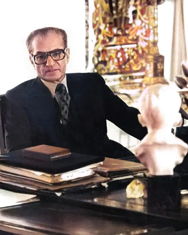 His Majesty, the Shah of Iran in a dark suit and glasses sits at a desk with books and a bust — a public domain photograph from The Shah of Iran archive documenting the Pahlavi era.