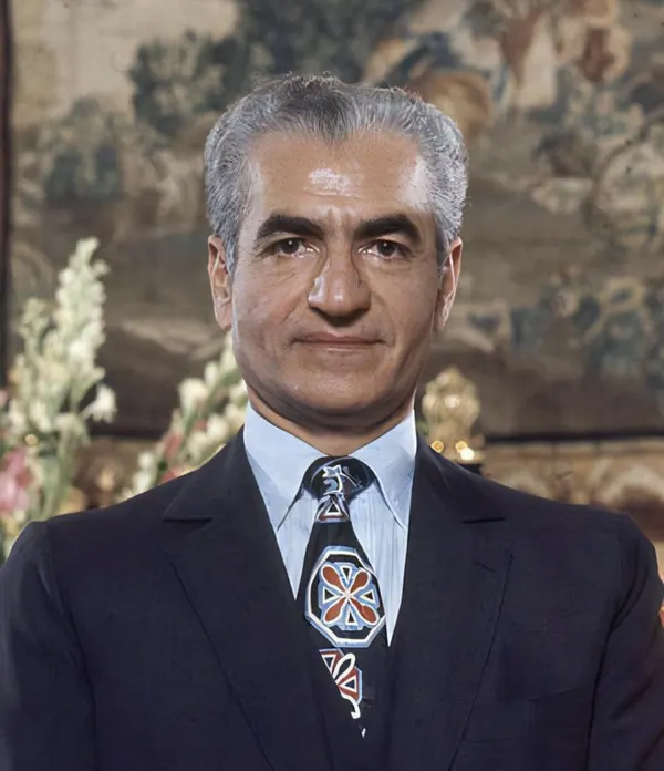 His Majesty, the Shah of Iran in a dark suit and patterned tie poses for a portrait against a tapestry — a public domain photograph from The Shah of Iran archive documenting the Pahlavi era.