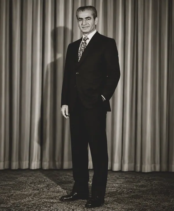 His Majesty, the Shah of Iran in a dark suit and patterned tie stands before a pleated curtain backdrop — a public domain photograph from The Shah of Iran archive documenting the Pahlavi era.