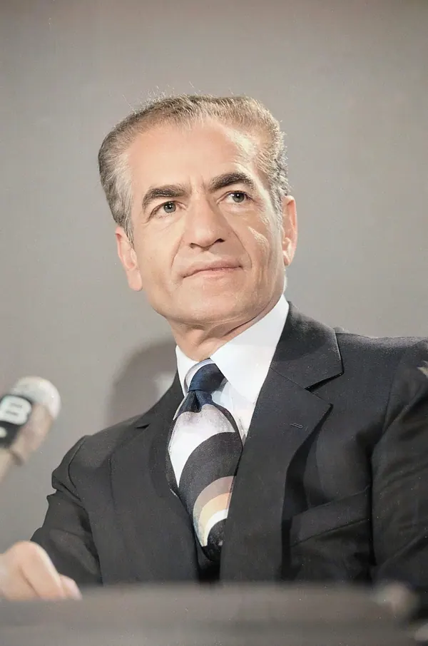 His Majesty, the Shah of Iran in a dark suit and striped tie looks slightly upward beside a microphone — a public domain photograph from The Shah of Iran archive documenting the Pahlavi era.
